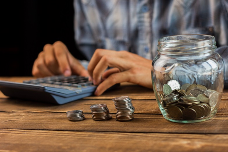 Person using a calculator with a jar of coins in the foreground.